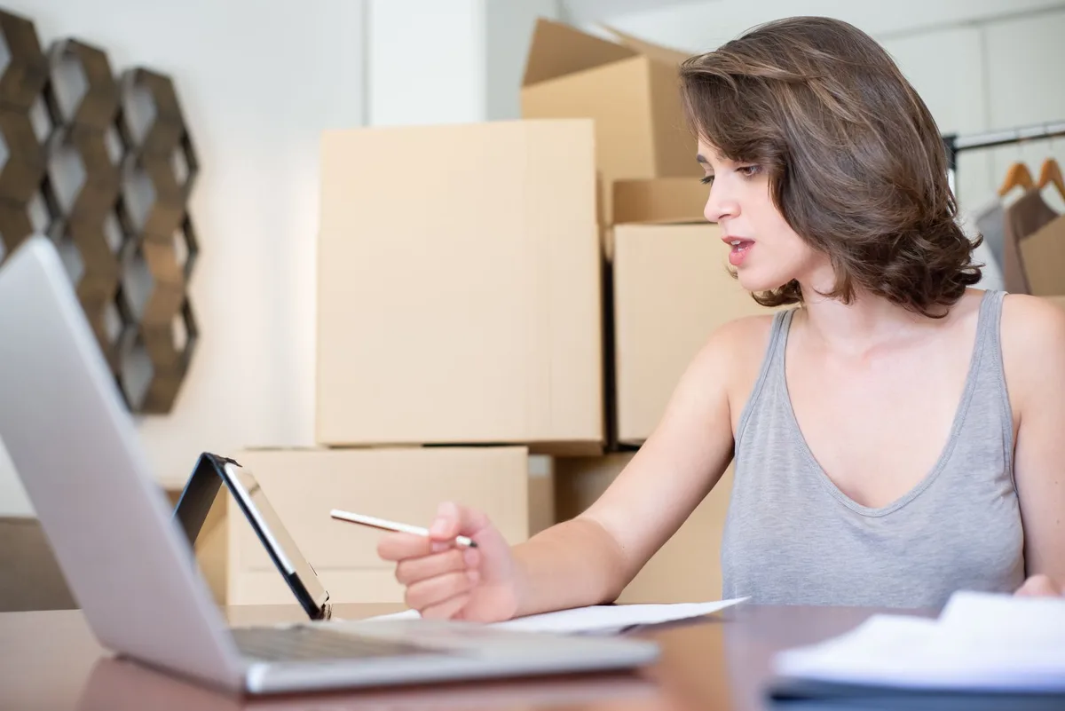 Online store owner reviewing orders and planning her affiliate program on a laptop, surrounded by packing boxes